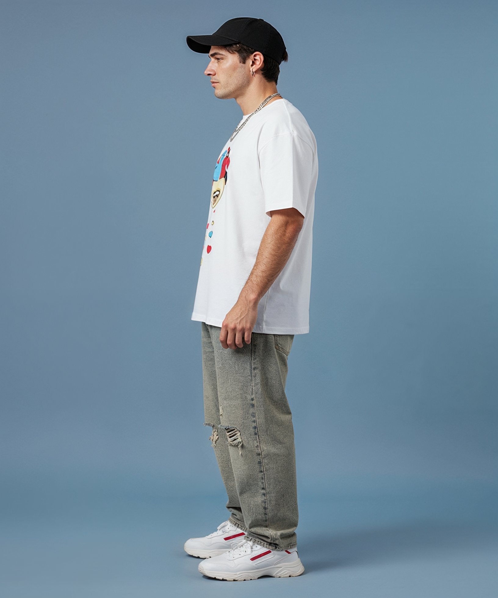male wearing a sweethearts white t-shirt with a colorful graphic design and ripped jeans on a blue background. side view