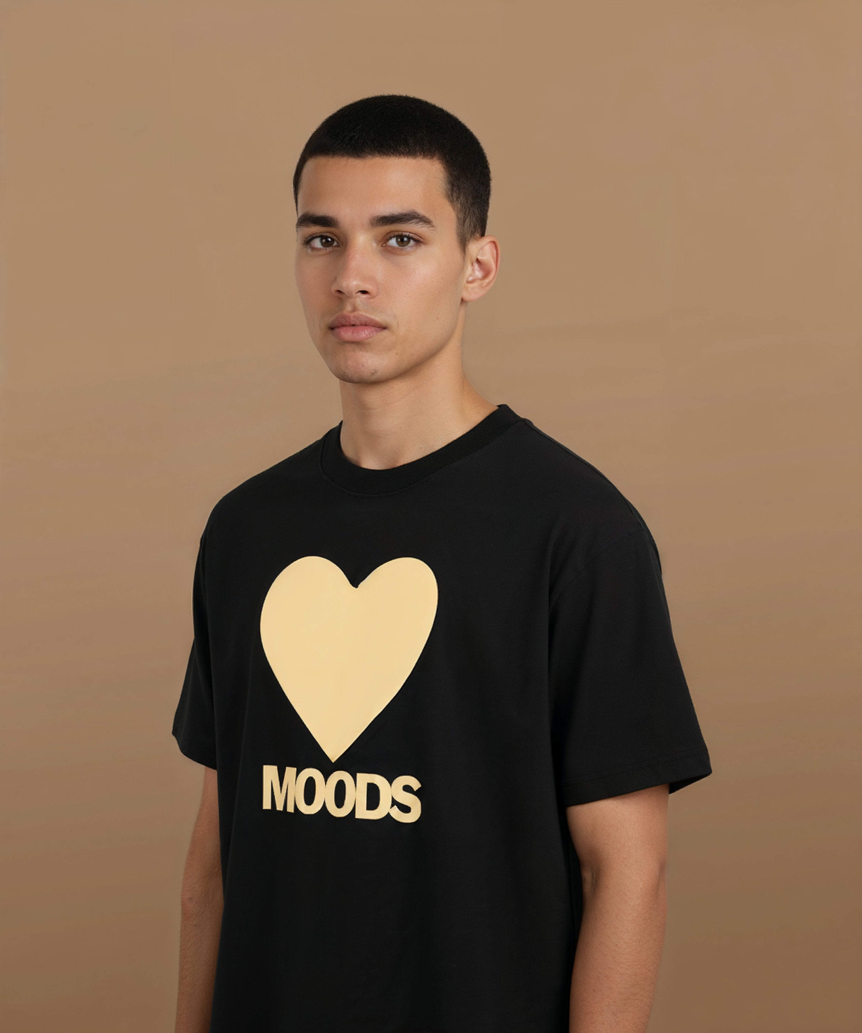 a male wearing a black sweethearts t-shirt with a gold heart and 'MOODS' text on a brown background, angled left