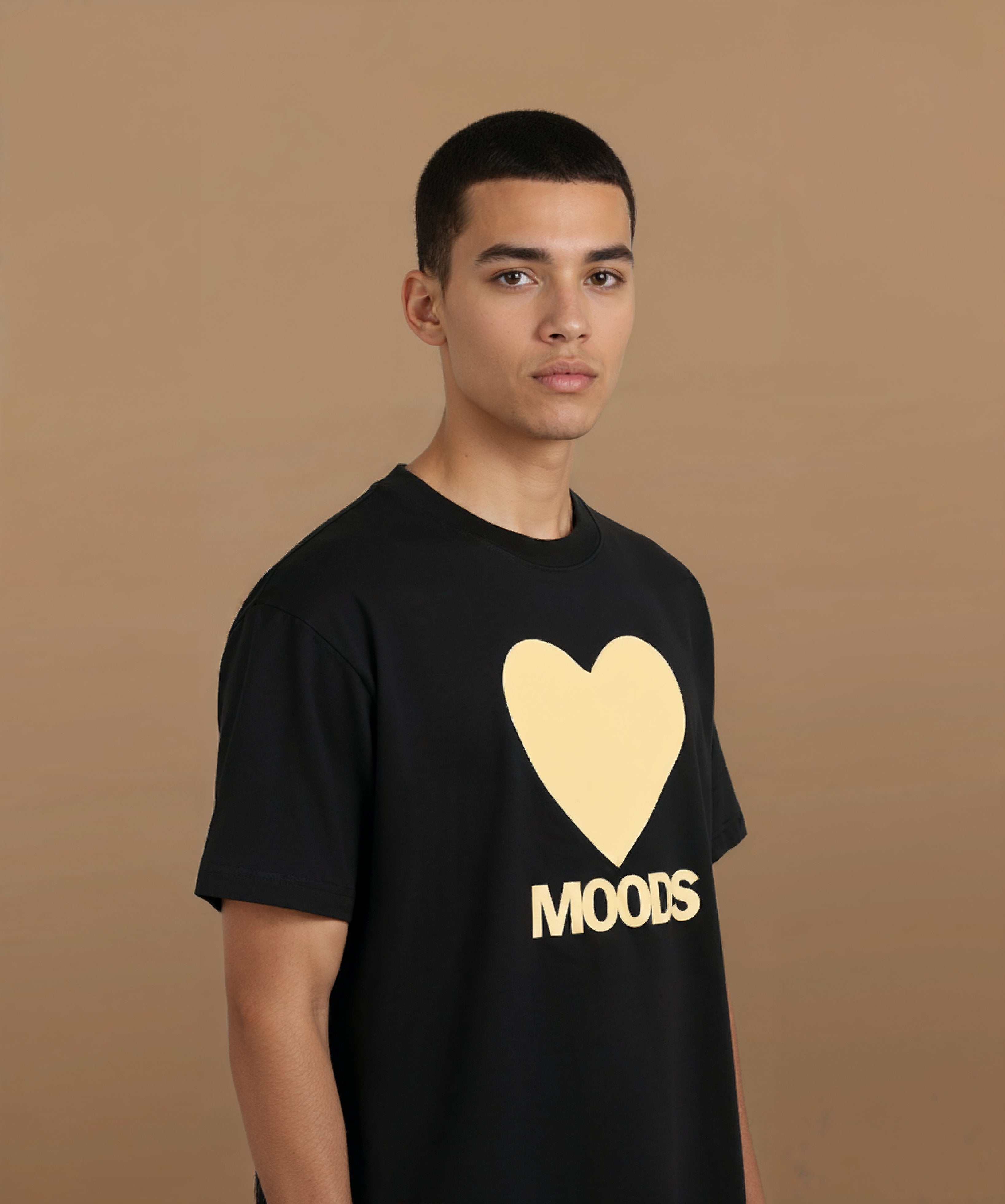 a male wearing a black sweethearts t-shirt with a gold heart and 'MOODS' text on a brown background, angled right