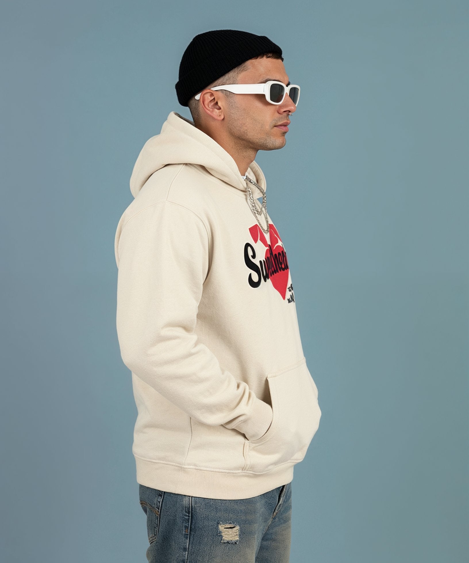 Man wearing a beige hoodie with a logo, black beanie, and white sunglasses on a gray background