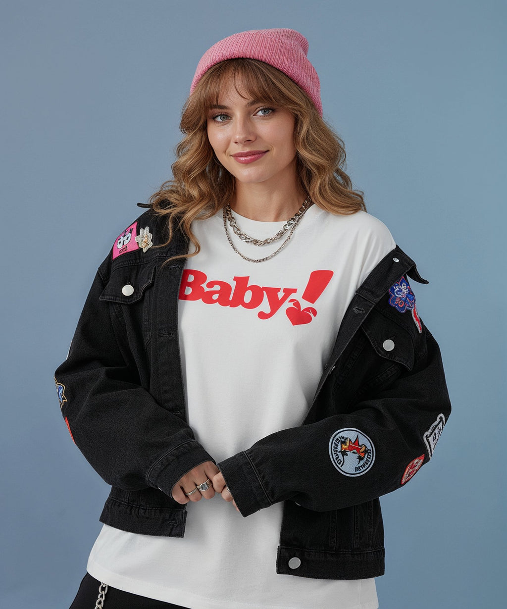 female wearing a black jacket with patches, white shirt with 'Baby!' print, and pink beanie against a gray background