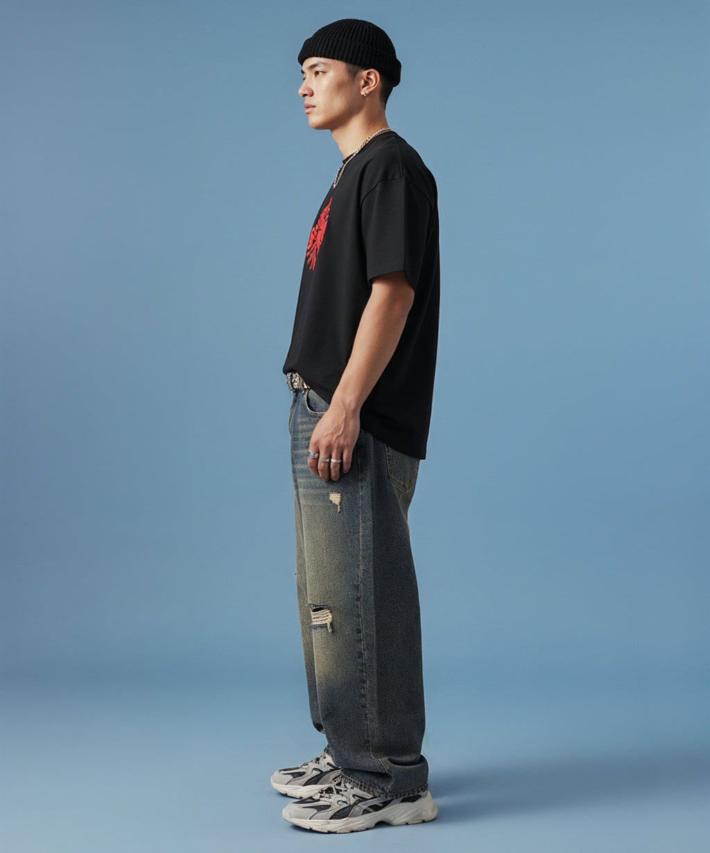 male wearing a black t-shirt with sweethearts red heart logo graphics and text in a circle and distressed jeans on a blue background, side view left