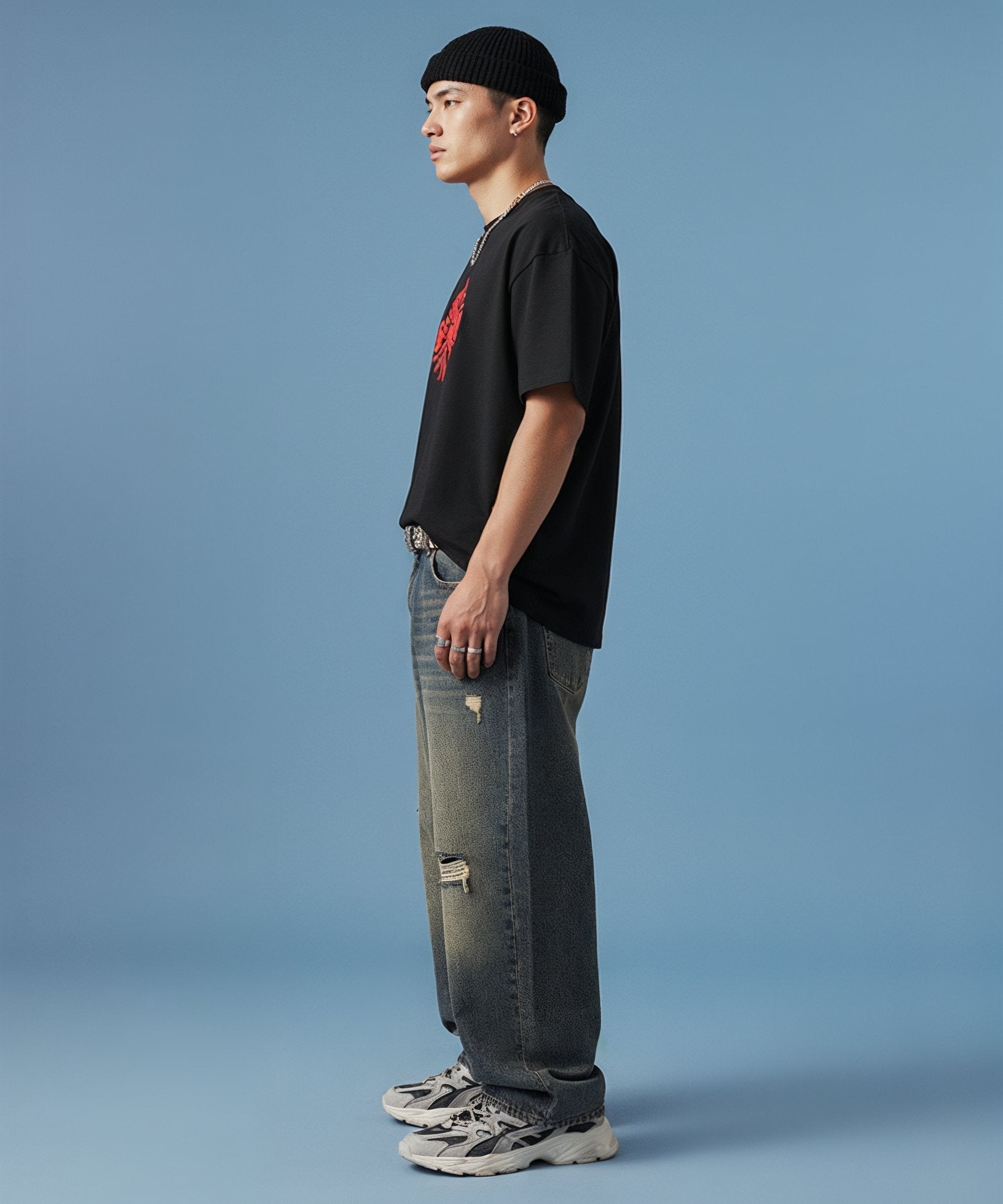 male wearing a black t-shirt with sweethearts red heart logo graphics and text in a circle and distressed jeans on a blue background, side view left