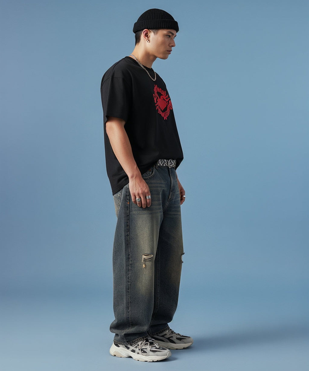 male wearing a black t-shirt with sweethearts red heart logo graphics and text in a circle and distressed jeans on a blue background, side view right