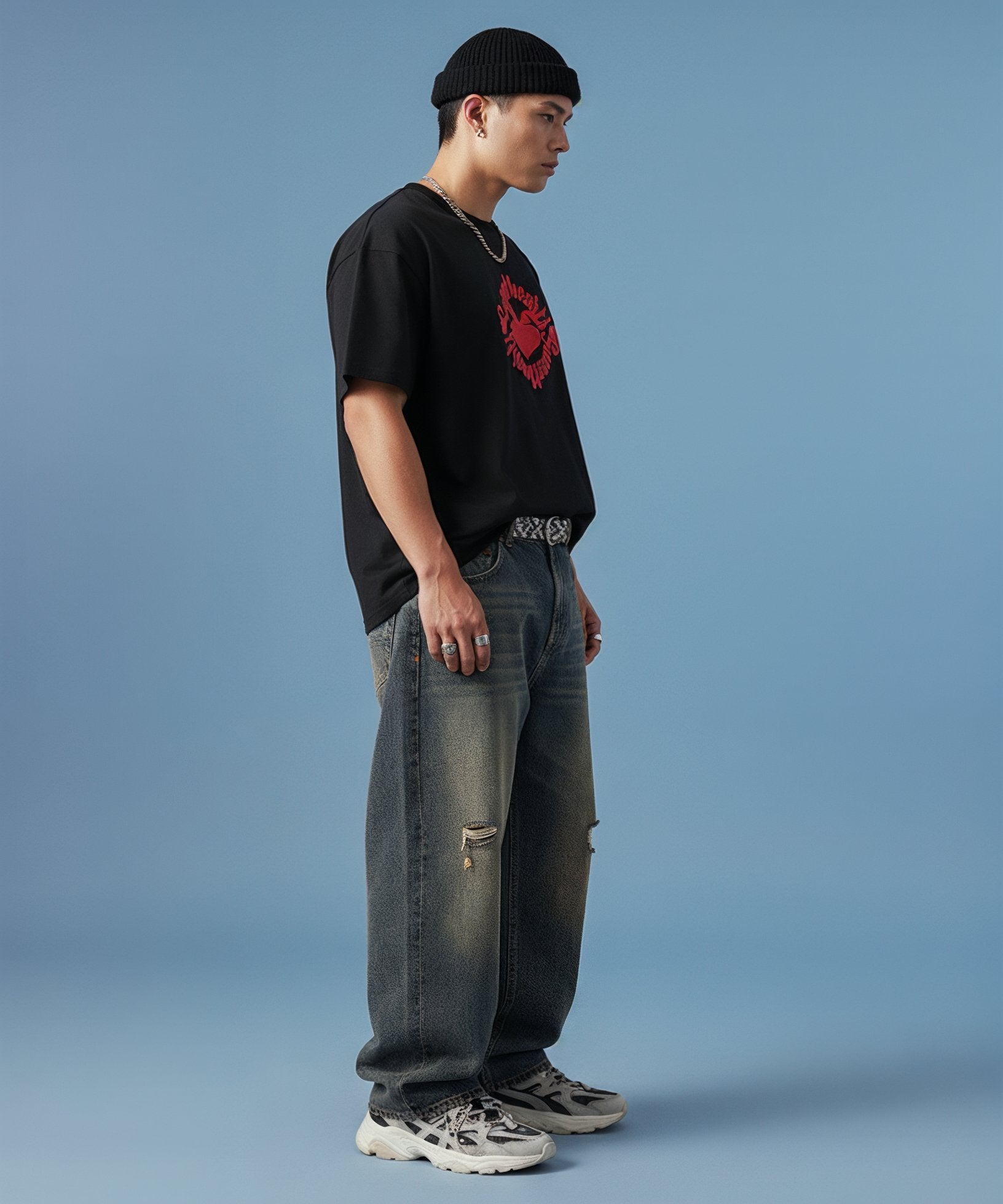 male wearing a black t-shirt with sweethearts red heart logo graphics and text in a circle and distressed jeans on a blue background, side view right