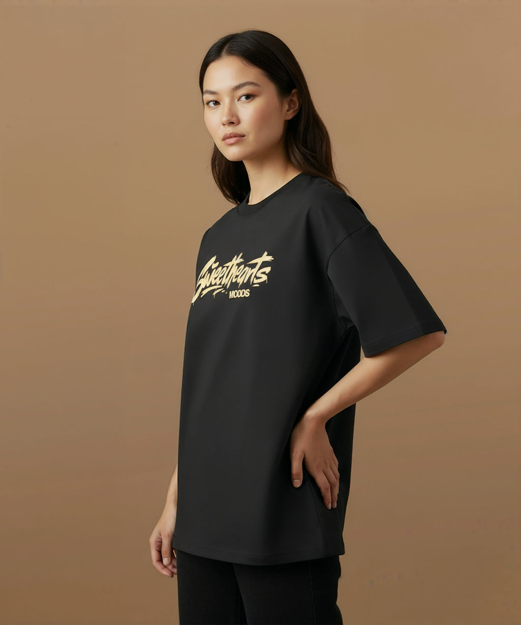 Feale model wearing a black t-shirt with 'Sweethearts' logo from moods collection,  graffiti text logo, on a brown background, angled left