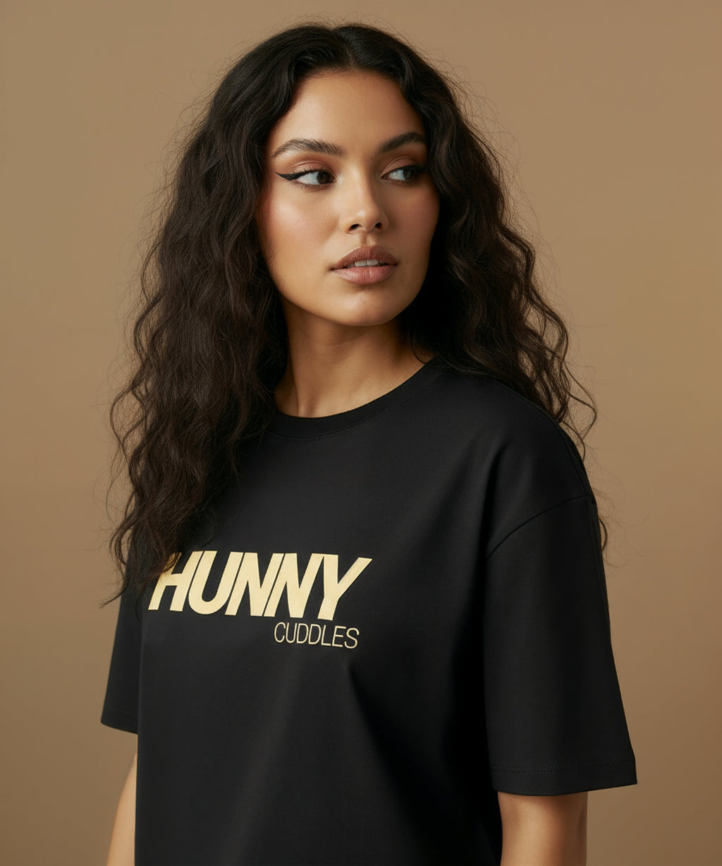 female model wearing a black t-shirt with 'HUNNY CUDDLES' text on a beige background, she is angled left