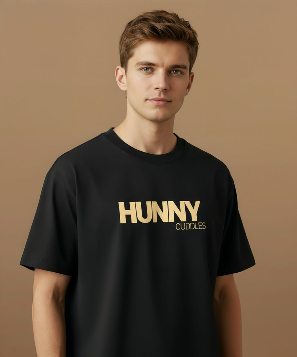 Male model wearing a black t-shirt with 'HUNNY CUDDLES' text on a beige background, angled right