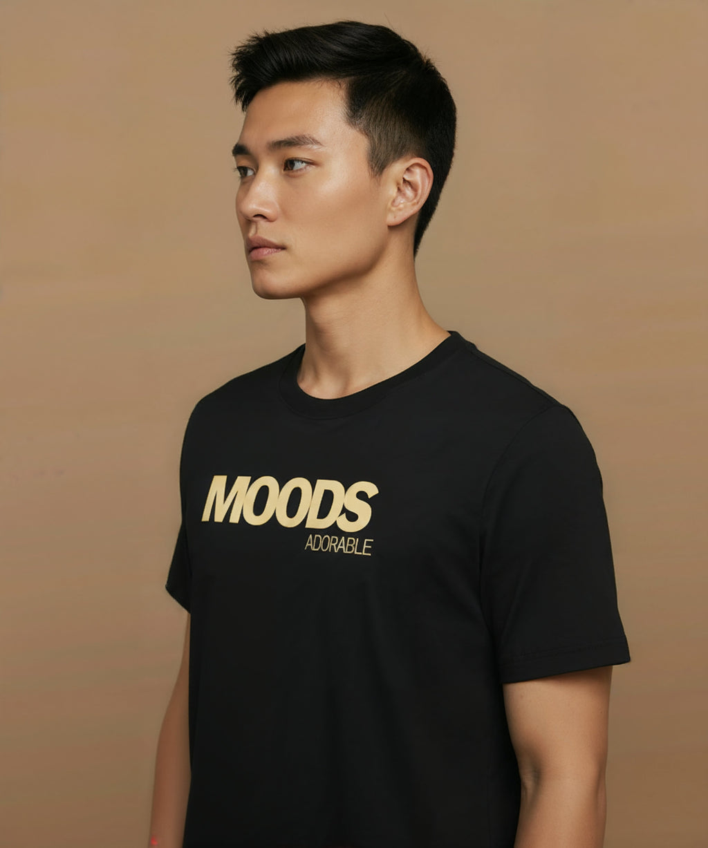 Man wearing a black t-shirt with 'MOODS', adorable, printed in gold on a brown background, angled left