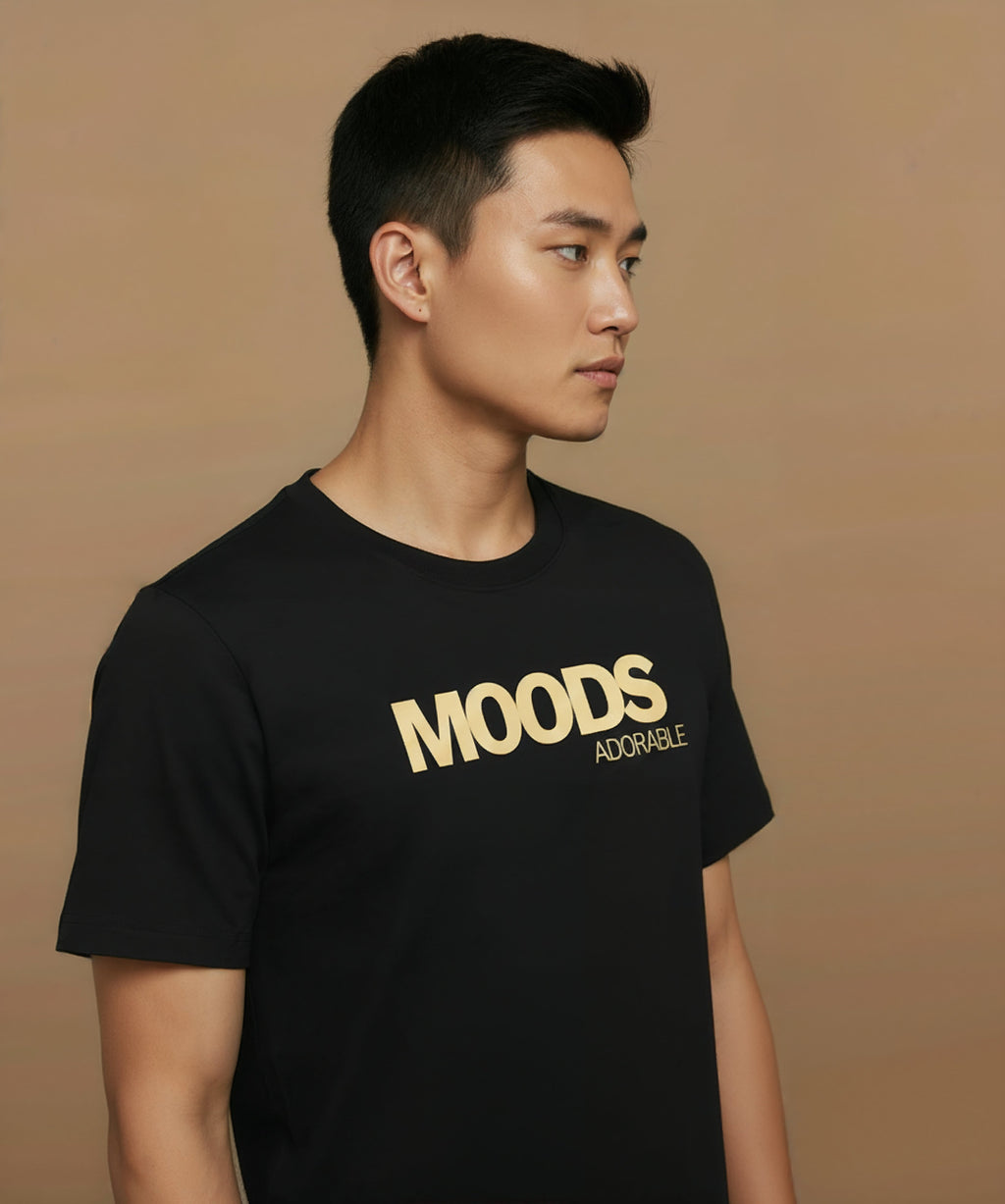 Man wearing a black t-shirt with 'MOODS', adorable, printed in gold on a brown background, angled right