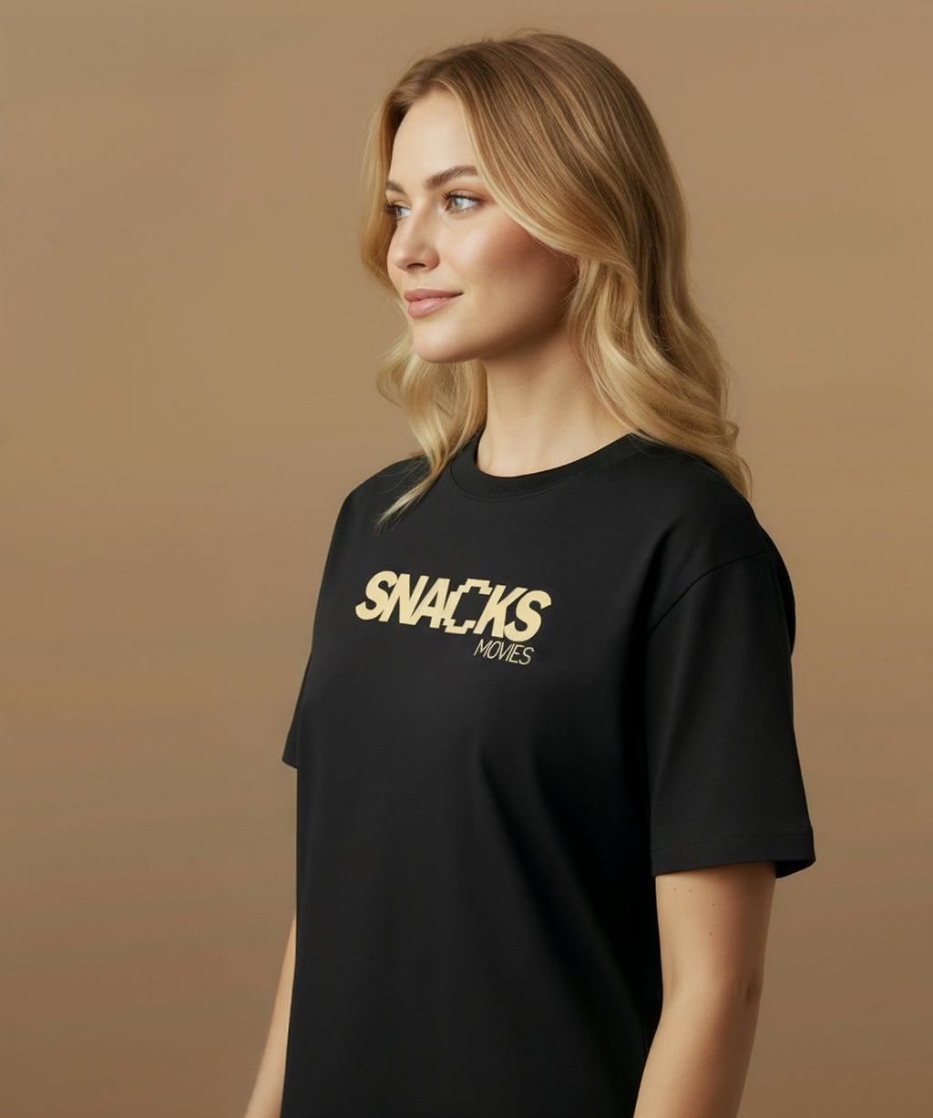 Female model wearing a black t-shirt with sweethearts brand 'SNACKS' printed in yellow from moods collection on a brown background. she is angled left