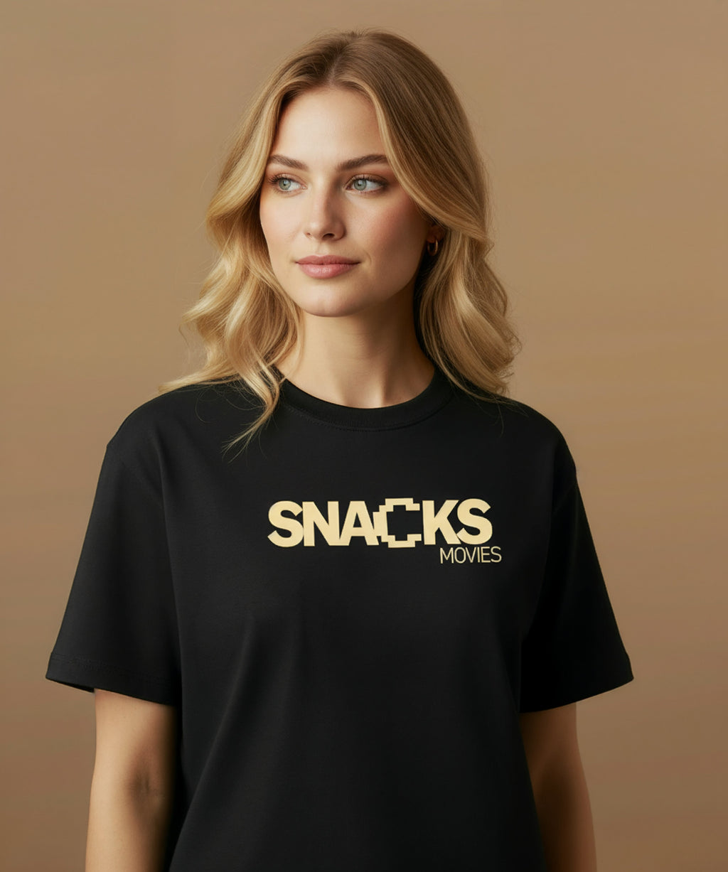 Female model wearing a black t-shirt with sweethearts brand 'SNACKS' printed in yellow from moods collection on a brown background. she is centered front
